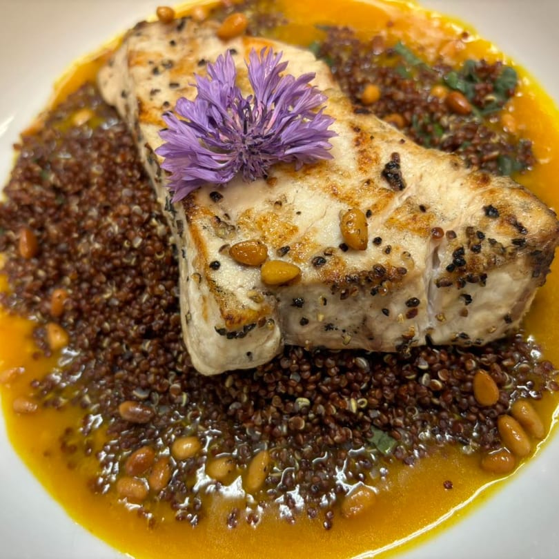Grilled fish with a purple flower garnish, served over quinoa, pine nuts, and a yellow sauce.