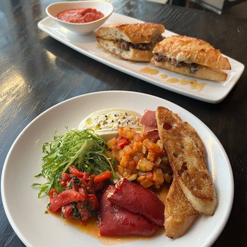 A gastropub spread featuring a charcuterie board and two steak sandwiches with a side of marinara.