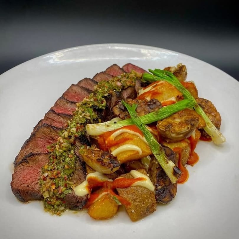 Sliced steak with green chimichurri, roasted potatoes, and grilled scallions on a white plate.